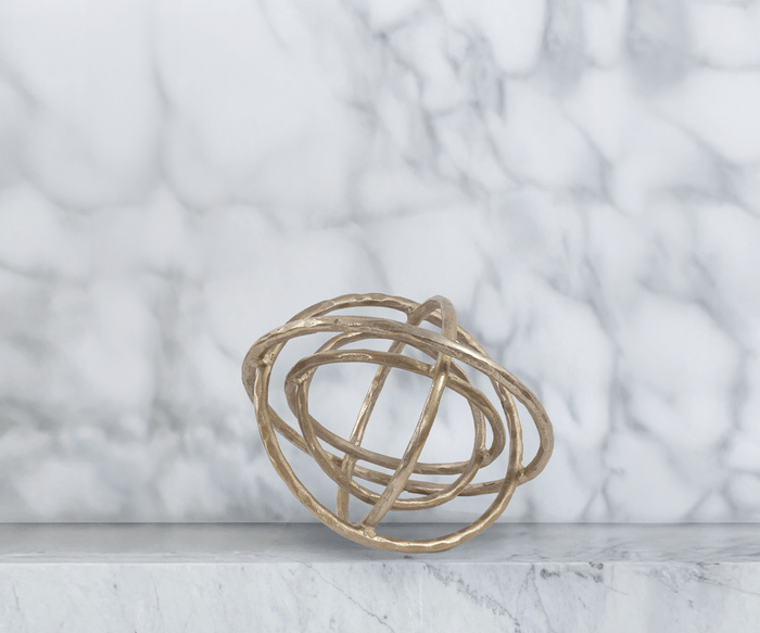 The Planetary Rings Sculpture, featuring interlocking antiqued brass aluminum rings, is displayed elegantly atop a white marble surface with a grey and white marble wall as the backdrop.