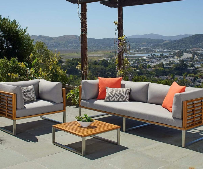 Modern outdoor patio featuring a sofa, an armchair, and the Playa Mar Outdoor Coffee Table. Gray cushions are accented with orange and patterned throw pillows, while the sleek teak finish beautifully complements the surrounding greenery and rolling hills under a clear sky.