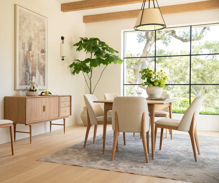 A bright dining room featuring light wood furniture, cream chairs, a large window with views of trees, a potted plant, and a sideboard adorned with fruit and decor. The Quiet Imprints Wall Art adds interest above the sideboard, while modern art elements and neutral tones create an airy, minimalist atmosphere.