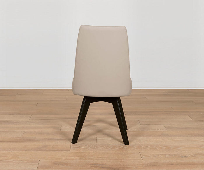 The Raynor Leather Swivel Dining Chair, upholstered in beige with a solid backrest and dark beech legs, sits on a light wood floor against a plain white wall, shown from the back.