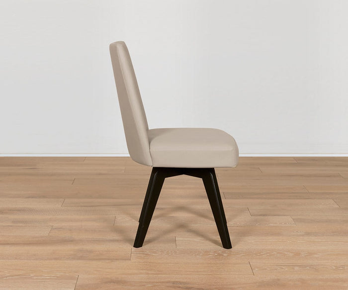 The Raynor Leather Swivel Dining Chair, shown in profile on a light wood floor against a plain white wall, features modern beige upholstery, a high back, and sleek black angled legs. This stylish chair combines comfort with contemporary design, making it an elegant addition to any dining space.