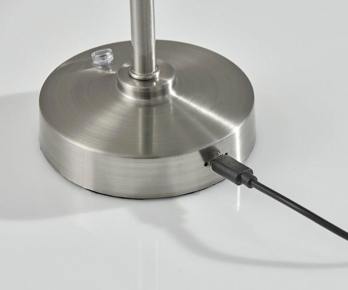 Close-up of the brushed steel base of the Ritchie Cordless LED Table Lamp, showing a power button on top and a black USB cable plugged into the side. The lamp sits on a white surface and features adjustable brightness for customized lighting.