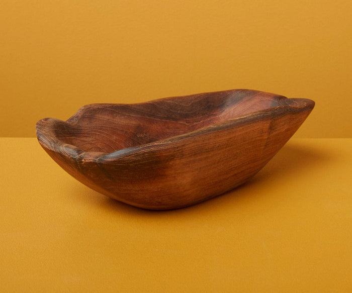 The Roscoe Teak Oval Dish, crafted from natural teak and featuring smooth, organic curves with a graceful silhouette, is displayed on a mustard yellow surface against a matching yellow background—an inviting earthy accent for any space.