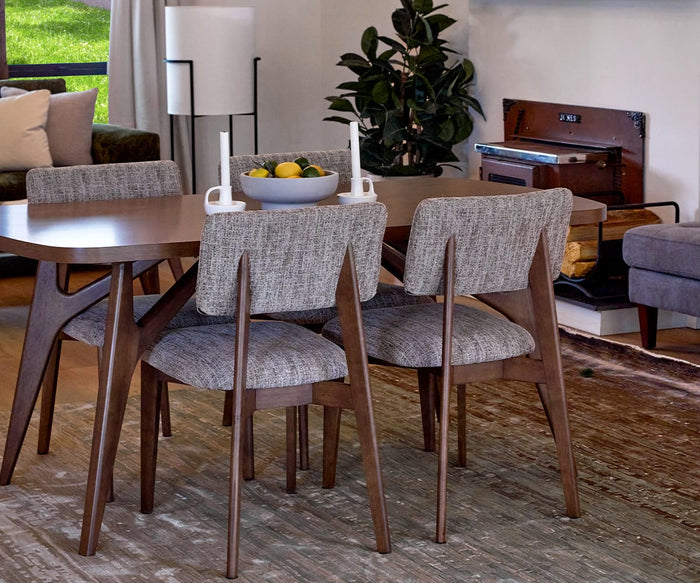 A modern dining area features four Saga Dining Chairs, a table holding a fruit bowl and candles, a gray sofa, a rug, a potted plant, and a window overlooking green grass outside.