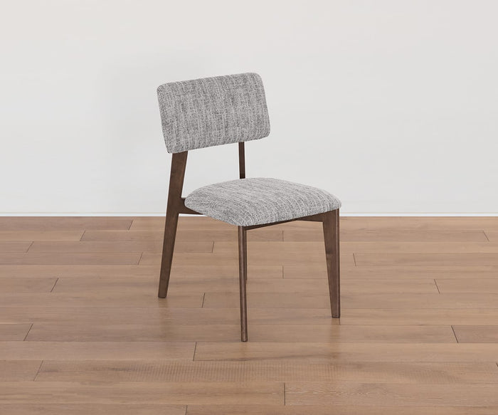 The Saga Dining Chair features wooden legs and a gray upholstered seat and backrest, shown here on a light brown wooden floor against a plain white wall.