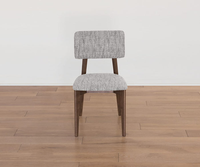 The Saga Dining Chair, crafted from wood and featuring a light gray upholstered seat and backrest, is placed on a light brown wooden floor against a plain white wall.