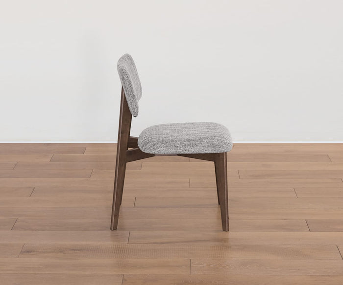 A side view of the Saga Dining Chair, showcasing its wooden frame with a cushioned seat and backrest upholstered in light gray fabric, placed on a light brown wooden floor against a plain white wall.