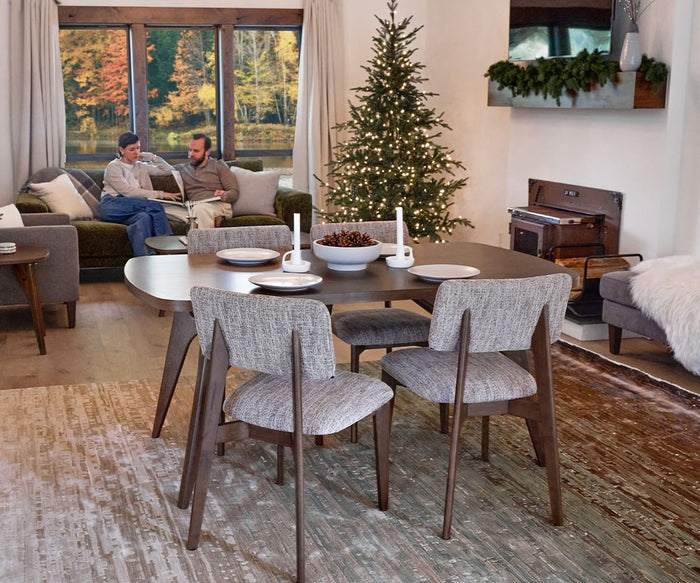 In a modern living and dining room, a set table is complemented by elegant Saga Dining Chairs, with a decorated Christmas tree, cozy seating, and two people conversing on a couch near a large window that offers a view of the forest.
