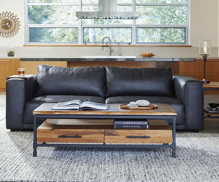 A modern living room features a black leather sofa, a wooden coffee table adorned with books and stones, and a gray rug. In the background, large kitchen windows frame vibrant greenery outside. The space is elegantly illuminated by the Samuel Table Lamp, whose antique brass base and sleek glass shade add both warmth and sophistication to the sophisticated setting.