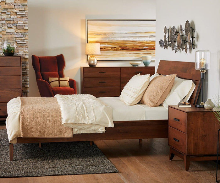 A cozy bedroom featuring a wooden bed with beige bedding, a matching nightstand and dresser, the Samuel Table Lamp—with its elegant glass shade and antique brass finish—wall art, metallic accents, and an orange armchair set in the corner on hardwood floors.