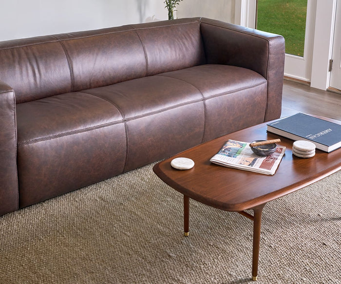 A brown leather sofa sits on a textured rug next to a wooden coffee table holding books, the Savina Round Marble Coasters - 4 Piece set, and a decorative bowl, near a window with a view of greenery.