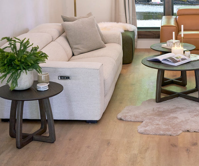 Modern living room with a cream sofa, wooden side tables adorned with Savina Round Marble Coasters - 4 Piece, candles, a plant, and books. The floor is light wood, and a soft beige rug rests near the tables. A large window lets in natural light.