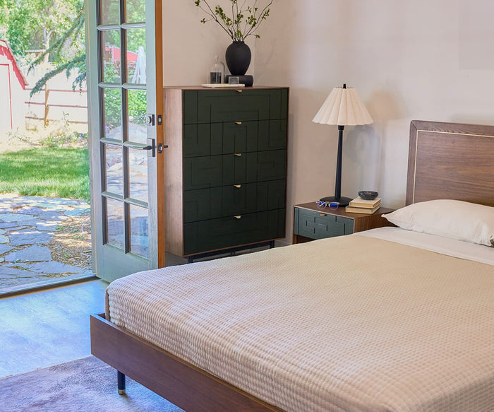 A tidy bedroom with a made bed, wooden furniture, and a green dresser features the Scallop Table Lamp, its scalloped shade and black wood base casting a warm glow. An open door reveals a sunny yard with grass and trees beyond.