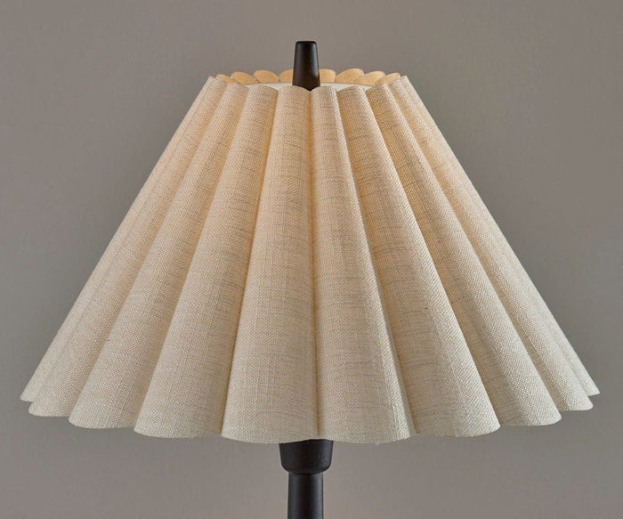 The Scallop Table Lamp features a beige pleated shade with a scalloped design perched on a dark base, set against a plain gray wall. Its textured, fan-like pleats gently diffuse the light, casting a warm glow that softens the room’s edges.