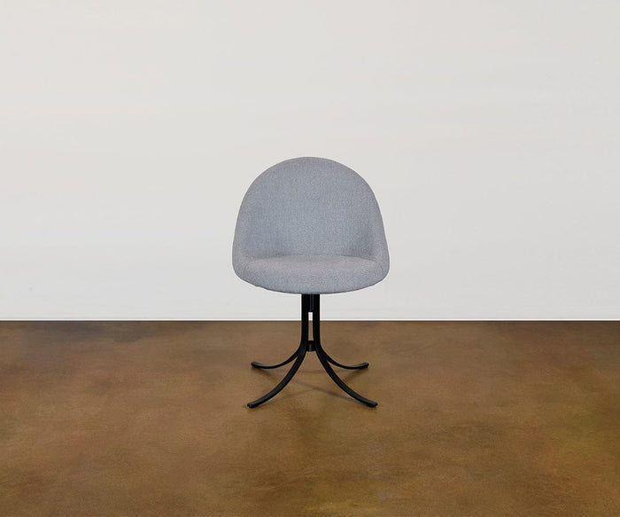 The Selene Dining Chair, with its modern gray upholstery, rounded backrest, and black metal legs, is placed on a brown floor against a plain white wall.