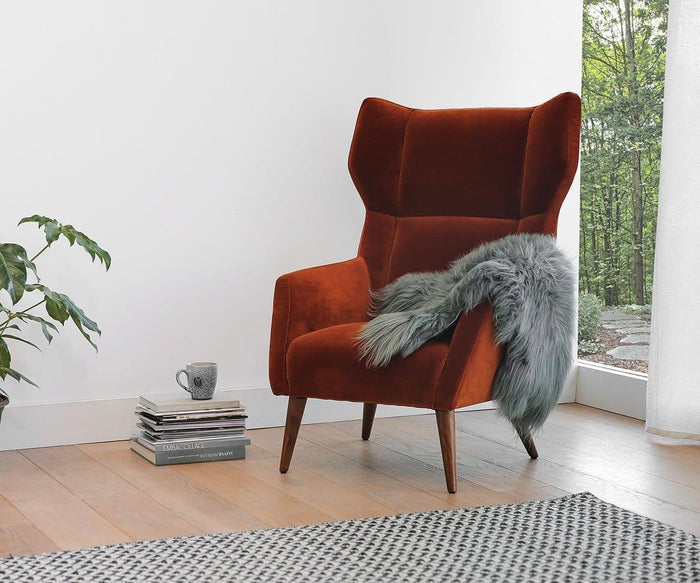A rust-colored armchair with a sumptuous Sheepskin Throw - Grey draped over it sits on a wooden floor next to a stack of books, a mug, a potted plant, and a window with greenery outside. A soft Scandinavian rug adds warmth and texture to the inviting space.