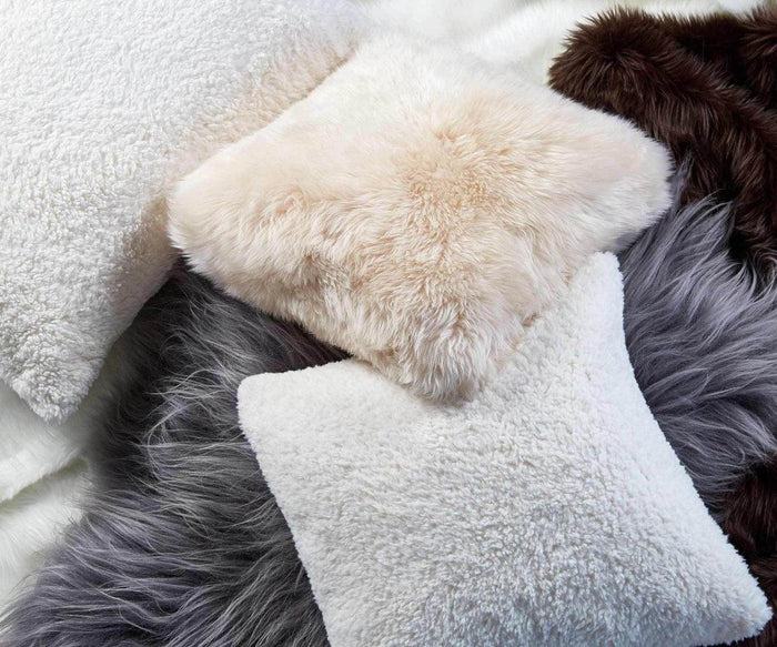 Four fluffy pillows in white, cream, and gray shades are arranged on matching furry blankets and an elegant Sheepskin Throw - Grey, creating a cozy and soft texture display.