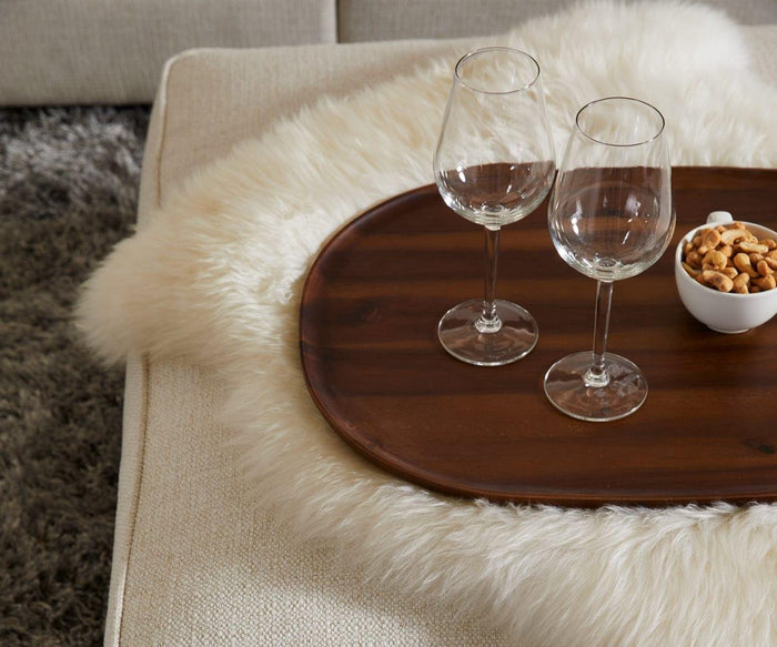 Two empty wine glasses and a small bowl of mixed nuts sit on a wooden tray placed on the Sheepskin Throw - Ivory, atop a beige sofa next to a gray rug—a true Scandinavian essential for cozy evenings in.