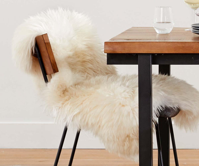 The Sheepskin Throw - Ivory, a fluffy white Scandinavian essential, is draped over a modern black metal chair beside a wooden dining table set with glassware, set against a light-colored wall and wooden floor.