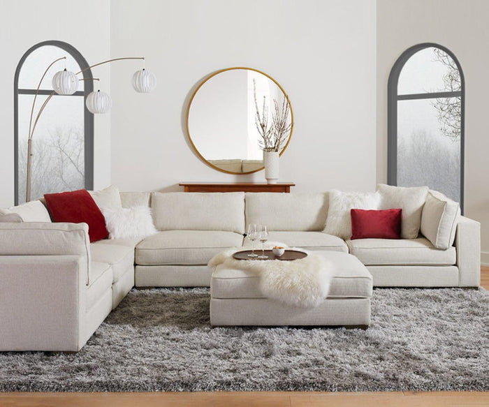 A modern living room features a large beige sectional sofa with red accent pillows, a round mirror, a floor lamp, and a shaggy gray rug crafted from soft New Zealand wool. At the center sits a round ottoman adorned with the luxurious Sheepskin Throw - Ivory and wine glasses. Two arched windows in the background complete this essential Scandinavian space.
