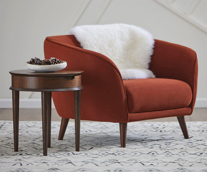 An upholstered red armchair with an ivory Sheepskin Throw sits beside a wooden side table topped with a bowl of pine cones, all resting on a patterned rug in this modern living room—a true Scandinavian essential.