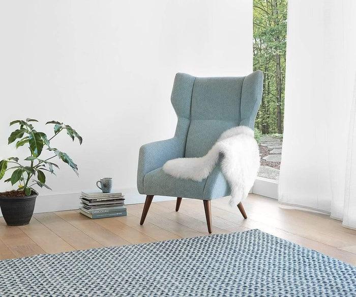 An ivory Sheepskin Throw drapes over a light blue armchair by the window, creating a true Scandinavian essential. Nearby, a potted plant, stacked books, and a mug complete the cozy setting. A blue patterned rug made of plush New Zealand wool covers part of the wooden floor.