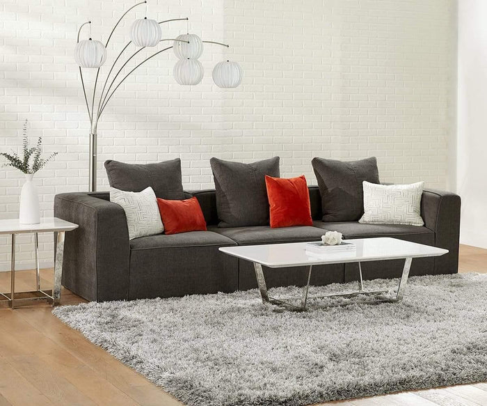 A modern living room with a dark gray sofa, red and white pillows, a white shag rug, and a sleek coffee table featuring stainless steel legs and a high gloss finish. Beside the seating area, there’s a Sieva End Table with a vase on top and a floor lamp with five round lights standing against the crisp white brick wall.