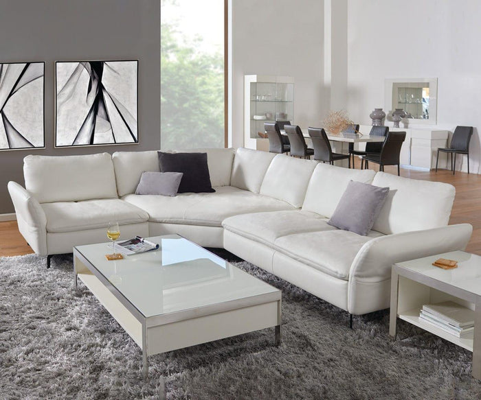 A modern living room features a white sectional sofa accented with gray throw pillows, a glass-top coffee table, a gray rug, and abstract wall art that conveys contemporary elegance. In the background, a dining area with a table and chairs adds to the open feel. The refined ambiance of the space is elevated by the dynamic artistry of the Silk Air II Oil Painting.