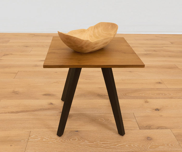 A small wooden side table with dark legs holds the Sindri Large Petal Bowl in mango wood, handcrafted in Thailand. The table sits on a light wood floor against a plain white wall.