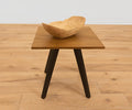 A small wooden side table with dark legs holds the Sindri Large Petal Bowl in mango wood, handcrafted in Thailand. The table sits on a light wood floor against a plain white wall.