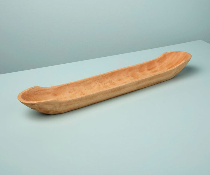 The Sindri Long Mango Wood Dish, handcrafted from sustainably sourced mango wood with rounded ends, sits on a light blue surface against a matching light blue wall.