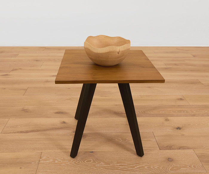 The Sindri Wave Edge Bowl, handcrafted from mango wood, sits on a small wooden table with black legs, placed on a light wood floor against a plain white wall.