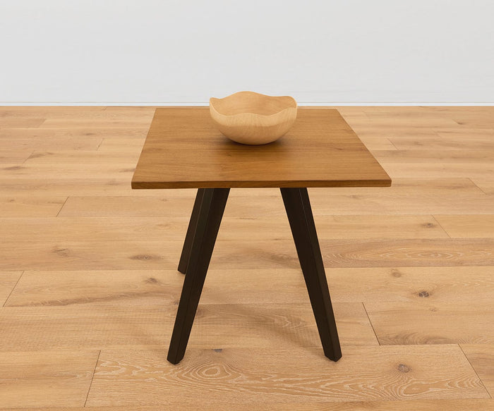 A small square wooden table with dark legs holds the Sindri Wave Edge Bowl, handcrafted from mango wood, set on a light wood floor against a plain white wall.