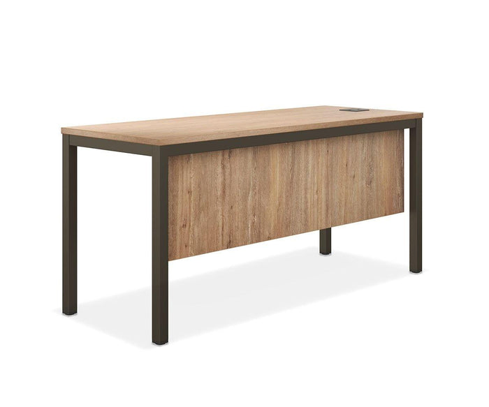 The Slater 63″ Desk features a rectangular tabletop with a light wood grain veneer and a matching front modesty panel, both supported by sturdy black metal legs. A visible cable grommet is located at the back right corner of the desk. The product is presented against a plain white background.