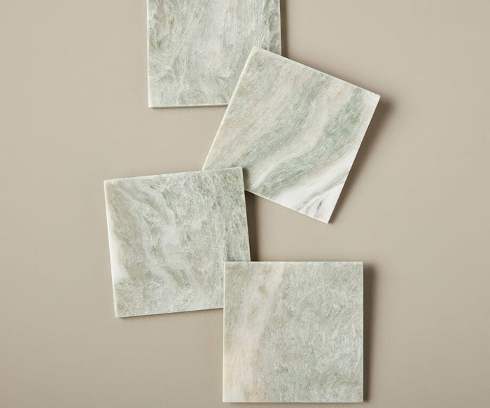Four Sora Lady Onyx Coasters, crafted from green marble and showcasing elegant swirls, are arranged in a slightly staggered, overlapping pattern on a beige background, creating a timeless aesthetic.