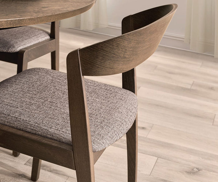 A close-up of the Soro Dining Chair featuring a curved backrest and a gray upholstered seat, placed next to a matching table on light-colored wood flooring. Soft natural light fills the room.