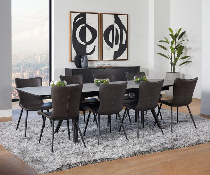 Modern dining room with a long table, eight Steele Dining Chairs arranged around it, a shaggy gray rug, abstract black and white art, a sideboard, a plant, and cityscape view through large windows.