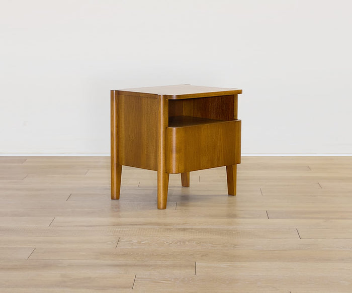 The Stefan Nightstand is a compact, Scandinavian-inspired piece in a mid-century modern style, featuring angled legs, an open shelf, and a single drawer. Finished in timber brown, it stands gracefully on a light wood floor against a plain white wall.