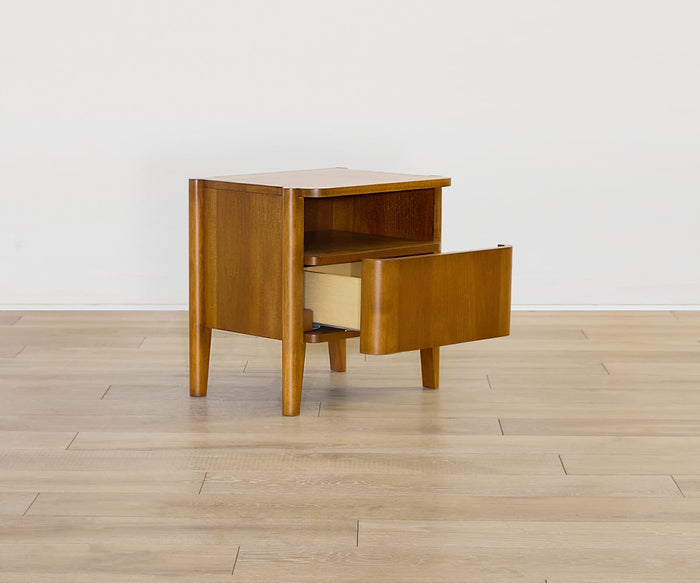 The Stefan Nightstand—a compact Scandinavian-style piece with four legs, one open shelf, and a single drawer slightly pulled out—rests on a light wood floor against a plain white wall.