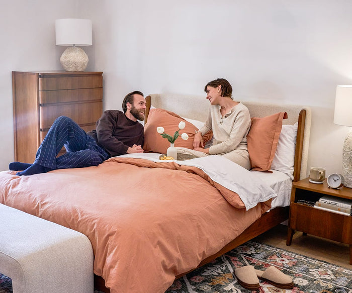 A man and woman sit on a neatly made bed with peach bedding, smiling and talking. The room features the Stefan Nightstand, along with a lamp, dresser, and cozy decor in a Scandinavian style.