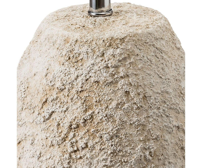 A close-up of the Stone Shores Table Lamp - Tall showcases its textured, rough beige stone-like base with a metallic rod extending from the top, set against a white background—evoking the understated elegance of modern home decor.