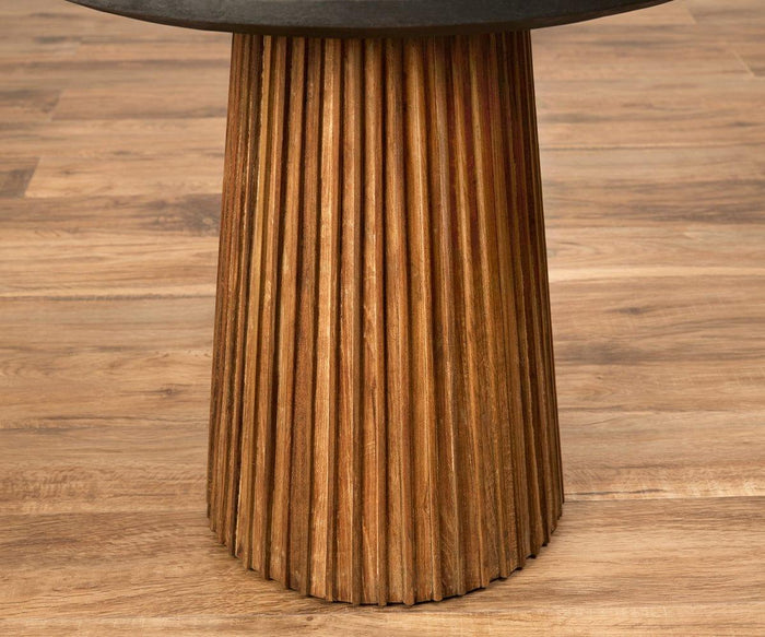 A Taavi 20″ Round End Table with a fluted, cylindrical base crafted from reclaimed teak and finished in a warm, natural tone sits on a light brown wood floor. The vertical grooves on the base highlight its artisan craftsmanship.
