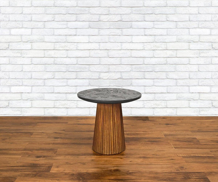The Taavi 20″ Round End Table, featuring a dark stone top and a wooden, tapered base crafted from reclaimed teak, sits on a wooden floor against a white brick wall backdrop. This piece captures the timeless appeal of mid-century modern design while beautifully combining sustainability with sophisticated style.