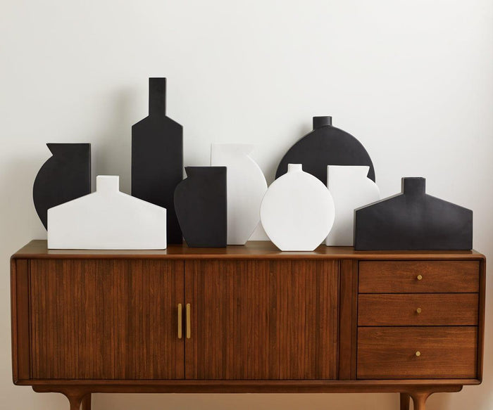 A wooden sideboard with brass handles displays eight geometric black and white vases, including a narrow vase and a contemporary Tall Silhouette Vase from the Tall Silhouette Vases collection—shaped like houses and bottles—against a plain white wall.