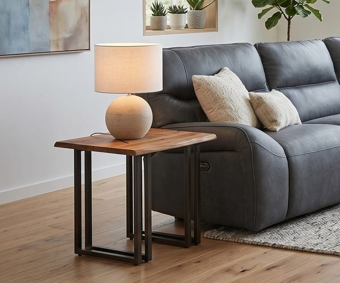 A wooden side table with the Textured Clay Lamp sits beside a gray leather couch with pillows. Plants, wall art, and a window letting in outdoor light complete the modern living room.