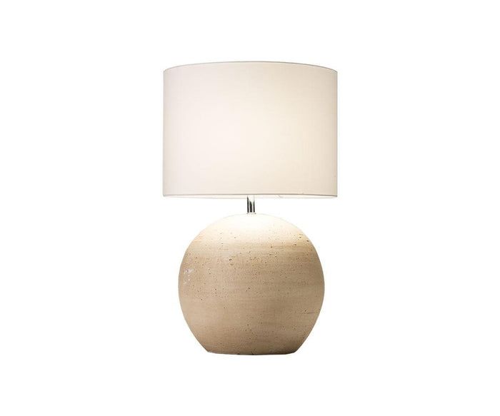 The Textured Clay Lamp features a round, beige base with a textured finish and a large, white fabric lampshade. It is elegantly centered on a plain white background.