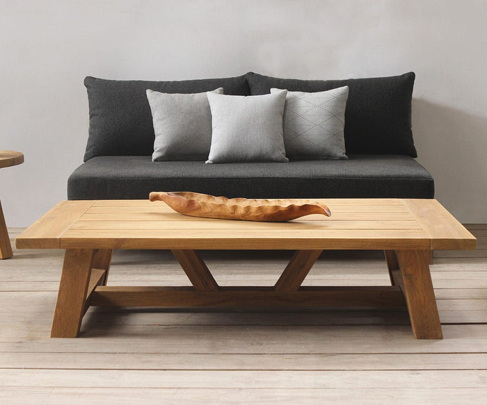 The Thorvid Outdoor Coffee Table, crafted from solid teak with a carved centerpiece and built for weather-resistant durability, sits in front of a dark gray sofa adorned with three decorative pillows in a minimalist living room, offering both style and resilience.