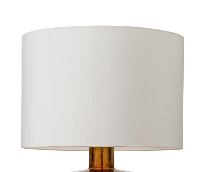 A white fabric shade rests atop the Translucent Glass Lamp - Amber, featuring a brownish-gold base and set against a plain white background to create an elegant look.
