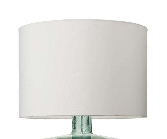 A close-up of the Translucent Glass Lamp featuring a white cylindrical fabric shade and a transparent recycled glass base, set against a plain white background.