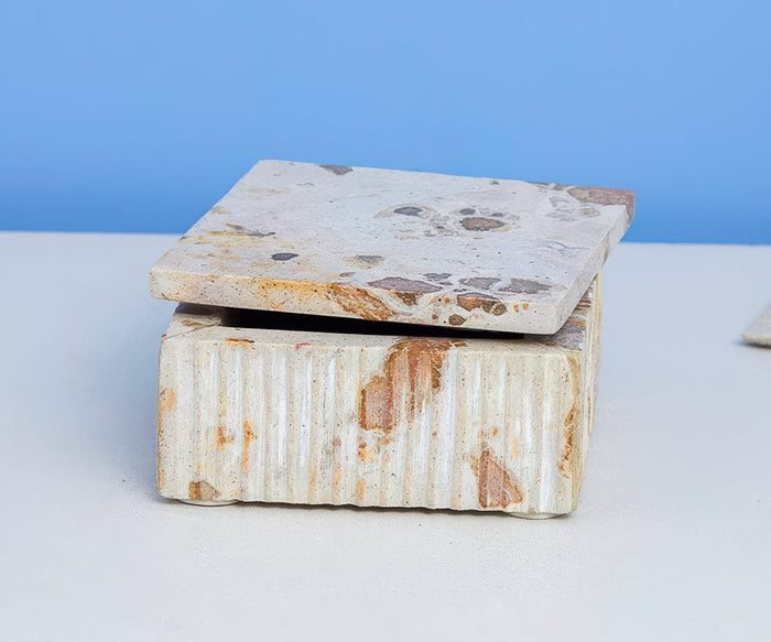 Two Tula Marble Boxes With Lid in White/Multi are stacked on a light surface, the bottom box featuring ridged detailing and the top box a smooth finish, creating a striking handcrafted decor piece against a blue background.